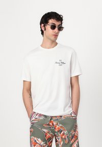 T-shirt en coton blanc avec un logo noir sur la poitrine gauche, associé à un short vert avec des motifs de feuilles orange et rose éclatants.