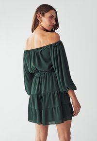 Grünes Off-Shoulder-Kleid mit puffigen Ärmeln, elastischem Bund und gestuften Rüschungen. Aus leichtem Stoff mit strukturierter Oberfläche gefertigt.