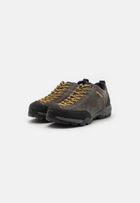 Pantofi de drumeție din suede gri, cu vârf din cauciuc negru și talpă durabilă. Caracteristici include șireturile galbene și toc întărit pentru susținere.