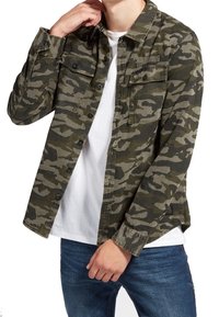 Camicia a motivo camouflage verde e nera con chiusura a bottoni, due tasche sul petto e maniche lunghe. Indossata sopra una maglietta bianca.