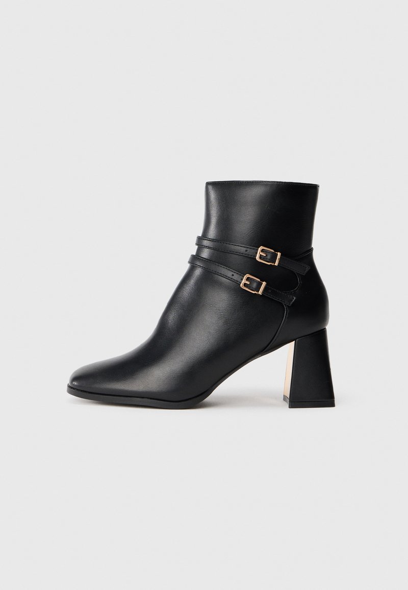 Bottine noire en cuir avec un bout pointu, dotée de deux sangles décoratives avec des boucles dorées et d'un talon épais. Texture lisse.