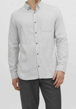 Homme portant une chemise à manches longues gris clair avec des boutons et une poche poitrine, ainsi qu'un pantalon gris foncé, debout devant un fond uni.