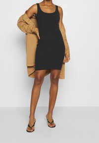 Femme portant une robe noire côtelée sans manches, des sandales noires à talons et un manteau beige tombant d'une épaule, sur un fond uni.