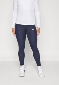 Marineblå leggings med tettsittende design, laget av stretchy stoff. Har hvitt Adidas-logo på nedre venstre lår. Brukt med hvit langermet topp og sko.
