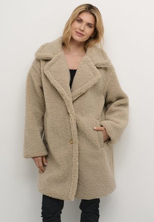 Beige fluffig kappa med en stor krage, knappstängning framtill, två sidofickor och ett mjukt strukturerat tyg.