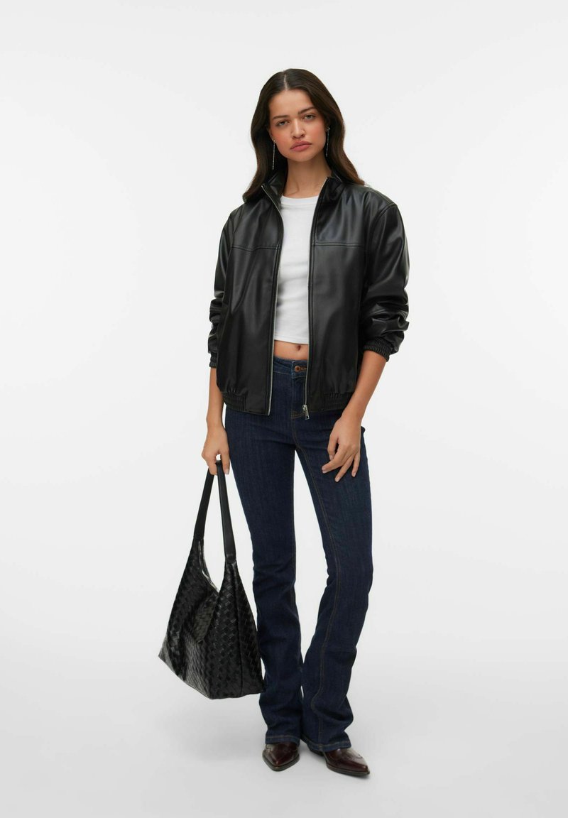 Femme portant une veste en cuir noire, un crop top blanc, un jean foncé et des chaussures pointues marron, tenant un grand sac cabas noir texturé.