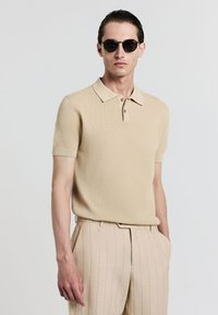 Beiger Strick-Polo mit strukturiertem Muster, kurzen Ärmeln und klassischen Kragen, kombiniert mit hellbeigen gestreiften Hosen.