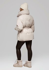 Nafoukaná béžová zimní bunda s vysokým límcem a páskem, kombinovaná s černými legínami a béžovými botami s kožešinovou podšívkou. Měkká, hladká textura.