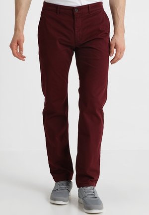 Man die nauwsluitende donkerrode broek en grijze sneakers draagt, staand tegen een effen witte achtergrond.