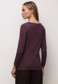 Maglia a maniche lunghe in viola intenso, realizzata in tessuto morbido. Presenta una scollatura rotonda e un design leggermente aderente con una texture liscia.
