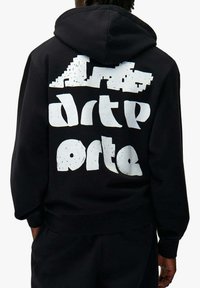 Person trägt einen schwarzen Kapuzenpullover mit großem weißem abstraktem Blocktext und grafischem Design auf der Rückseite.