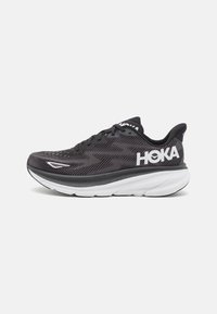 Scarpe da corsa nere e grigie con una tomaia in rete, una suola intermedia bianca imbottita e un prominente logo bianco "HOKA" sul lato.