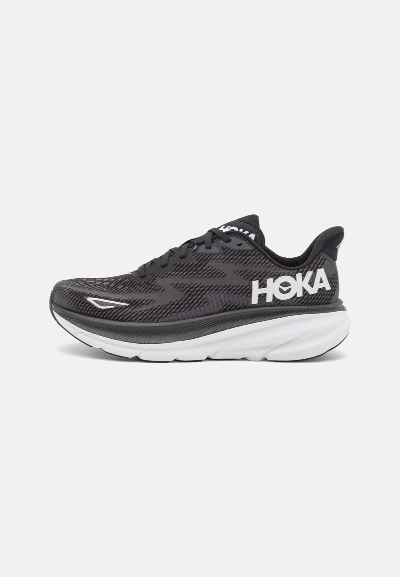 Scarpe da corsa nere e grigie con una tomaia in rete, una suola intermedia bianca imbottita e un prominente logo bianco "HOKA" sul lato.