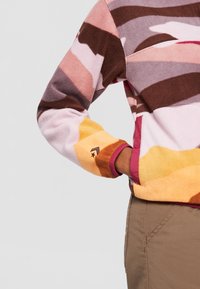 Persona vistiendo una chaqueta de forro polar a rayas multicolores con la mano en el bolsillo lateral, combinada con pantalones marrones sobre un fondo sencillo.