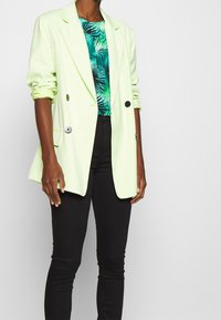 Blazer verde chiaro con due bottoni neri, caratterizzato da un design sartoriale. Abbinato a un top con motivo tropicale e jeans neri a vestibilità slim.