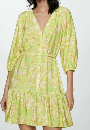 Femme portant une robe à motif floral vert et beige avec des boutons sur le devant et des manches bouffantes trois-quarts.