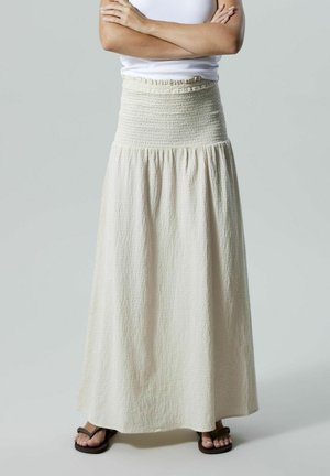 Jupe maxi couleur crème avec une taille smockée, un tissu texturé et une coupe fluide, associée à des sandales marron.