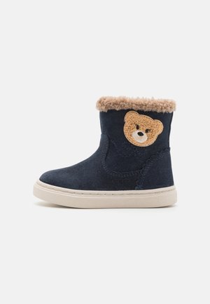 Mørkeblå semsket støvler med en beige, fluffy krage, med en teksturert bjørneapplikasjon på siden og en hvit såle. Kort ankelhøyde design.