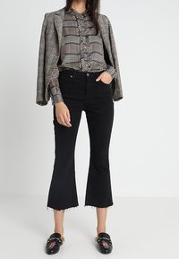 Jeans neri a zampa d'elefante con orli grezzi, abbinati a una camicia a quadri marrone e un blazer grigio. Mocassini neri con ricami floreali completano il look.