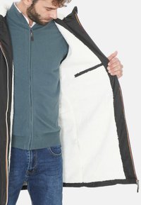 Giacca nera con fodera soffice bianca, chiusura con zipper e tasca interna. Pullover blu a strato intermedio visibile sotto. Jeans in denim.