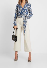 Kvinna iklädd blå blommig omslagsblus, off-white vida cropped byxor, högklackade sko i ormskinn, hållande en svart handväska.