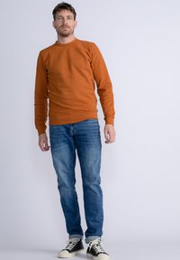Sudadera naranja con cuello redondo, con mangas largas y puños de canalé, combinada con jeans de mezclilla azul y zapatillas negras de caña alta.