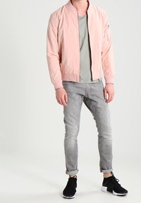 Cazadora bomber de color rosa claro hecha de tela suave, con cremallera frontal, bolsillos laterales y puños acanalados, combinada con vaqueros grises y zapatillas negras.