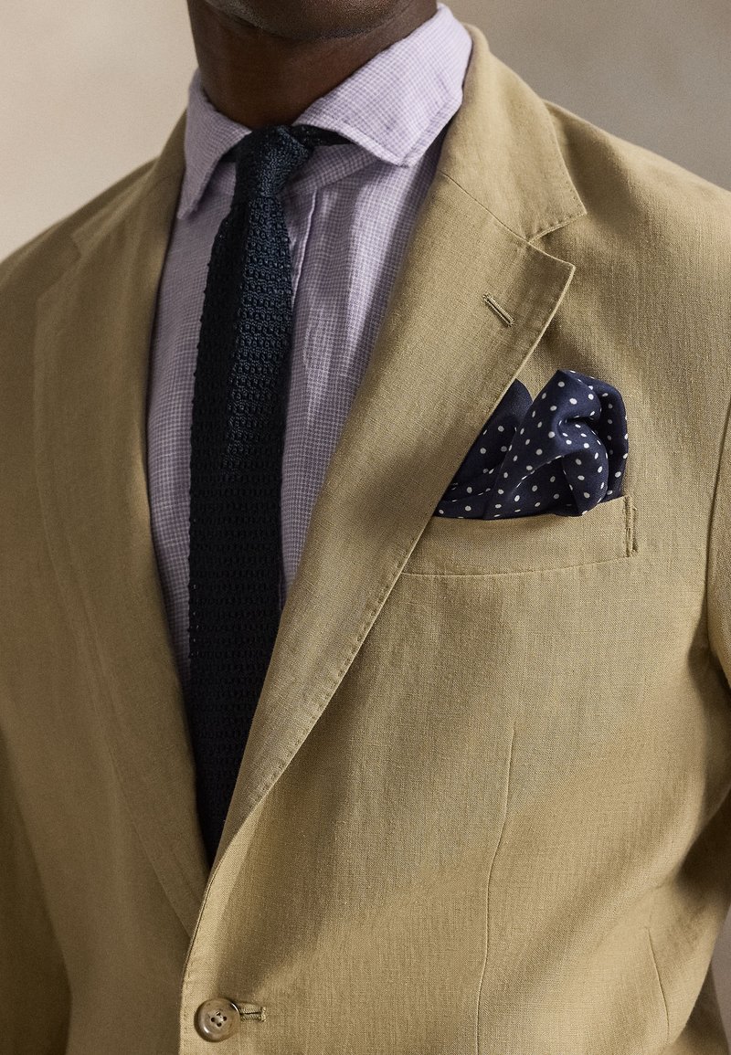 Light tan linen blazer with a single button, paired with a lilac checkered shirt and a navy polka dot pocket square. Textured black tie.