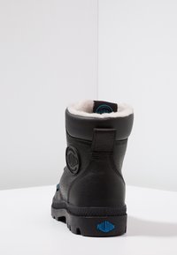 Stivale alla caviglia in pelle nera con finitura testurizzata, fodera crema e un logo blu circolare sul lato. Presenta una suola in gomma robusta.