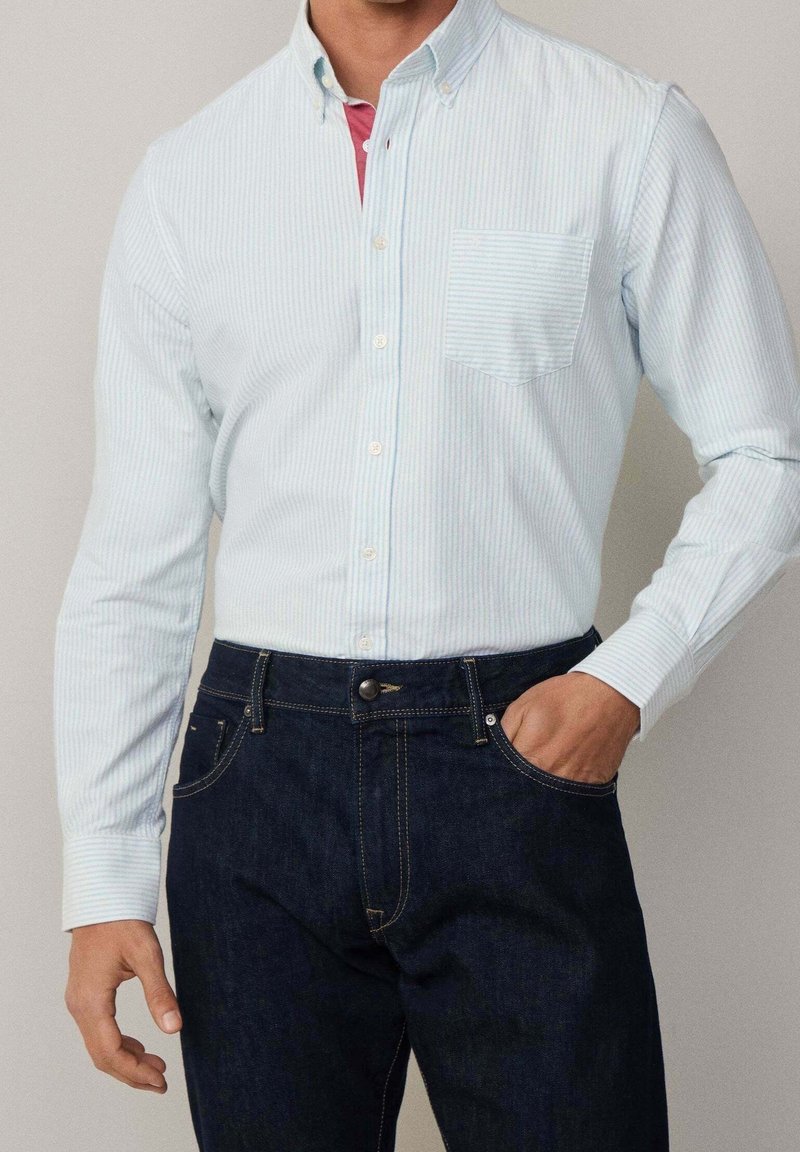 Homme portant une chemise rayée bleu clair avec poche poitrine, rentrée dans un jean bleu foncé, une main dans la poche.