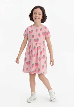 Mlada deklica se smeji, oblečena v roza obleko z jagodnimi vzorci in belimi visokimi supergami, stoji ob belem ozadju.