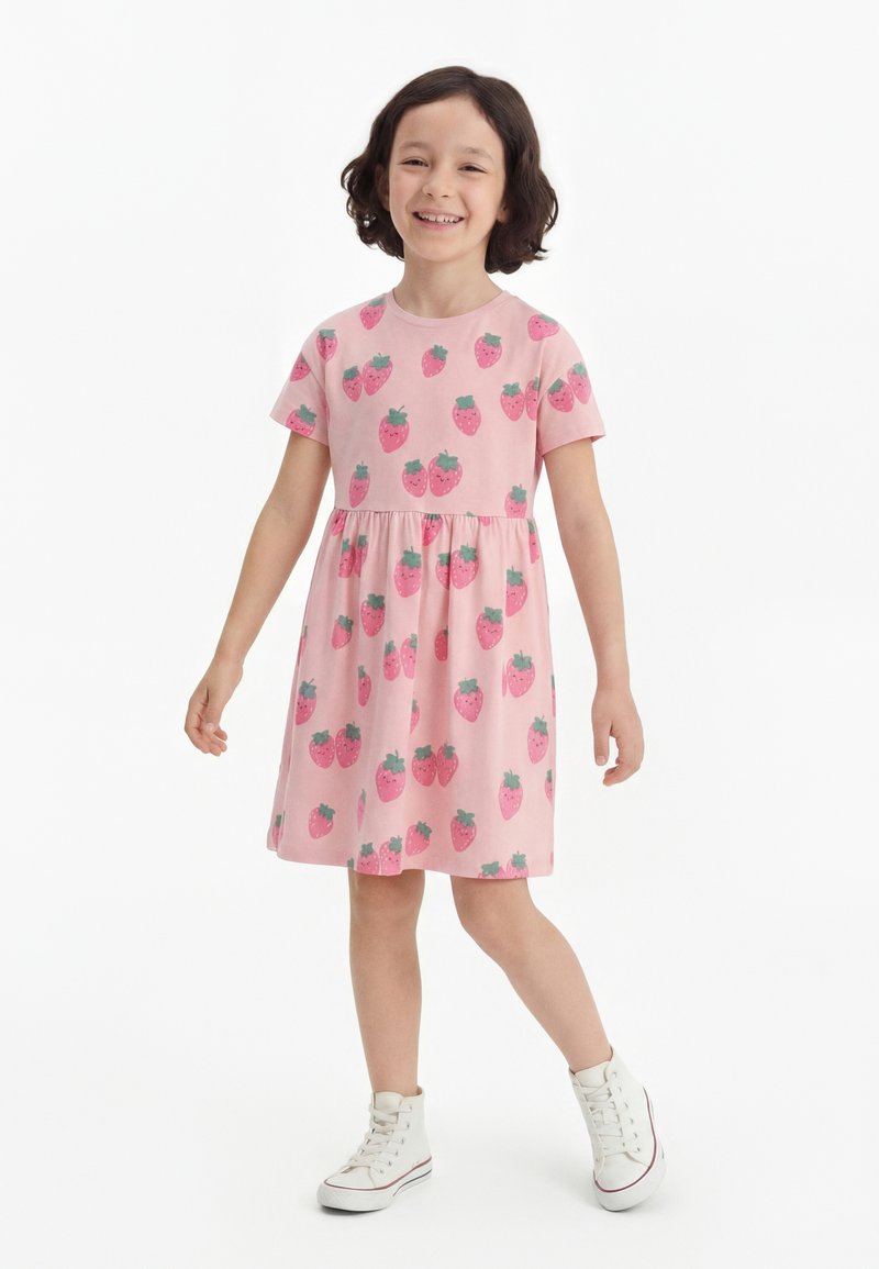 Νεαρή κοπέλα που χαμογελά, φοράει ένα ροζ φόρεμα με στάμπες φράουλας και λευκά ψηλά αθλητικά παπούτσια, στέκεται μπροστά σε λευκό φόντο.