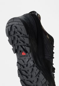 Μαύρο και καμουφλαριστό ορειβατικό παπούτσι σε γωνία για να δείξει την ανθεκτική σόλα με βαθιές εγκοπές και το κόκκινο λογότυπο Salomon στο κάτω μέρος.