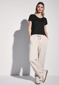 Donna in piedi con le mani in tasca, indossando una maglietta nera a maniche corte, pantaloni beige a gamba larga e sneakers bianche con suole marroni.