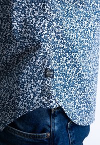 Gros plan de l’ourlet d’une chemise fleurie bleu et blanc sur un jean en denim bleu foncé avec des coutures visibles et une petite étiquette noire.