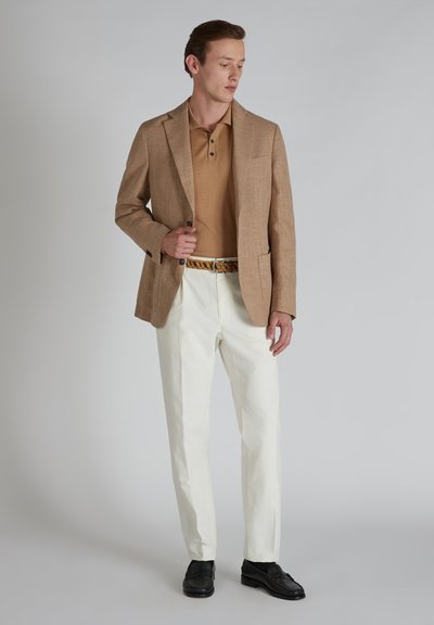 Blazer texturizado marrón claro sobre un polo beige, combinado con pantalones blancos y zapatos negros. El modelo está de pie frente a un fondo neutro.
