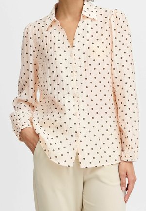 Vrouw die een lichtroze blouse met lange mouwen en zwarte polkadots draagt, met beige broek, één hand in de zak.