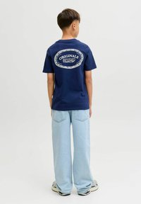 Navyblauwe katoenen t-shirt met een witte grafiek en "ORIGINALS STUDIO" op de achterkant. Gepaard met lichtblauwe wijdemaat jeans en sneakers.