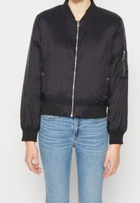 Veste bomber noire avec fermeture éclair à l'avant, poignets et ourlet côtelés, et poche latérale zippée, fabriquée en matériau lisse et brillant.