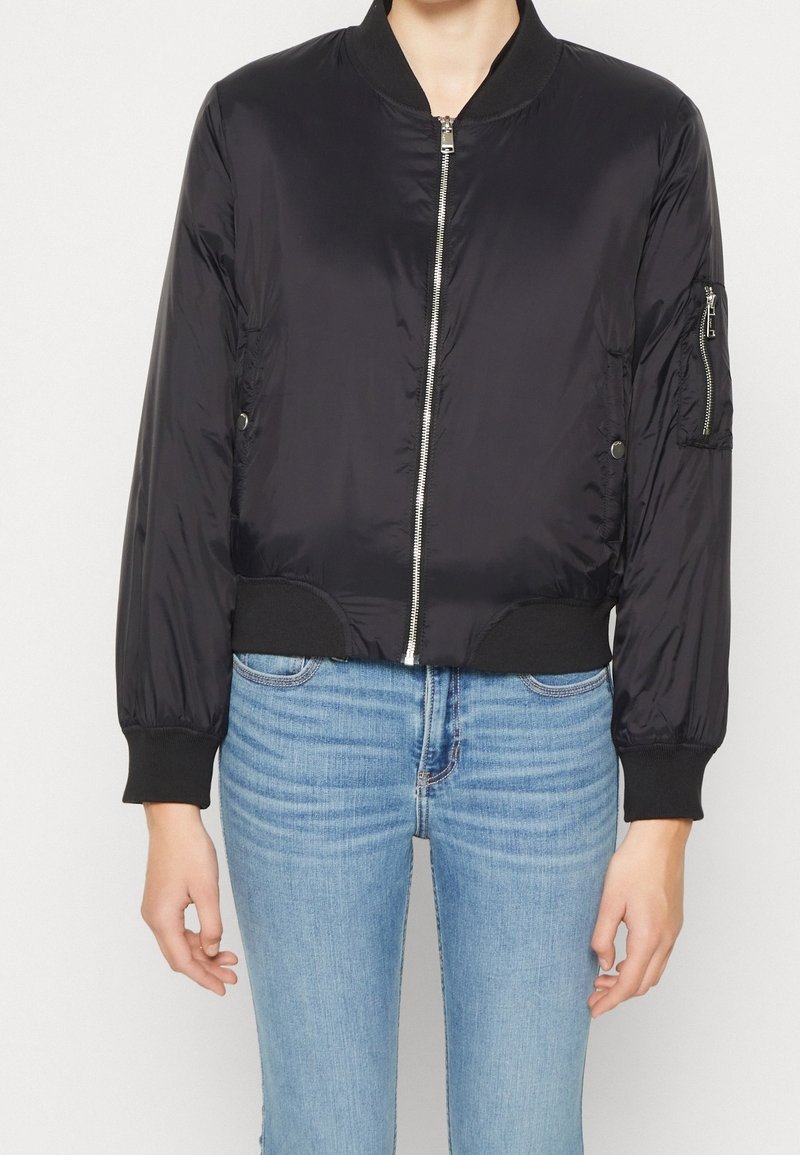 Veste bomber noire avec fermeture éclair à l'avant, poignets et ourlet côtelés, et poche latérale zippée, fabriquée en matériau lisse et brillant.