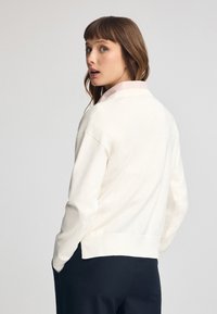 Maglione color crema con una vestibilità rilassata, orlo a coste e maniche arrotolate, indossato sopra una camicia colletto a quadri leggeri rosa.