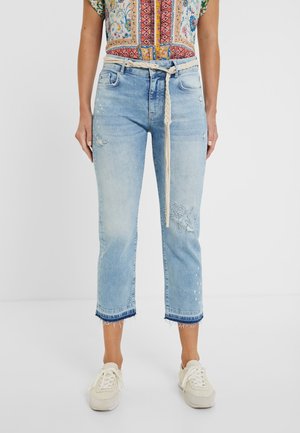 Femme portant un jean court bleu clair avec des ourlets effilochés, une broderie florale, une ceinture tressée beige, un haut à motifs colorés et des baskets blanches.