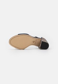 Scarpa con tacco alto di colore blu navy, con un accento dorato intrecciato sul tacco e una suola liscia beige, caratterizzata da un design elegante e punta arrotondata.