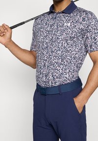 Blommig polo skjorta med marinblå bas, prydd med rosa och vita blommor. Kombination med marinblå byxor och ett brett vävt bälte. Golfklubba hålls över axeln.