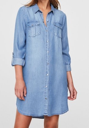 Robe chemise en denim bleu clair avec un devant boutonné, deux poches poitrine et des manches retroussées. Texture douce avec une coupe décontractée.