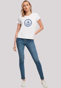Weißes T-Shirt mit einem blauen kreisförmigen Grafikdesign, das ein Segelboot und Pfeile zeigt, kombiniert mit blauen Jeans und weißen Sneakers.