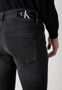 Jeans de mezclilla negra con una textura suave, que cuentan con un bolsillo trasero y una etiqueta de Calvin Klein Jeans en la cintura.
