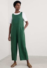 Žalias lininis šokinėjimas su plačiomis kojomis, turintis apvalų kaklą ir šonines kišenes. Derinamas su baltu marškinėliu ir rudomis diržinėmis basutėmis.