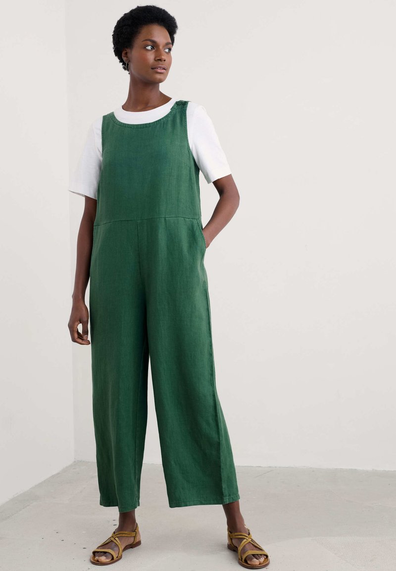 Žalias lininis šokinėjimas su plačiomis kojomis, turintis apvalų kaklą ir šonines kišenes. Derinamas su baltu marškinėliu ir rudomis diržinėmis basutėmis.