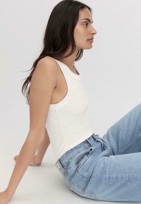 Weißes, geripptes Tanktop mit weiten Armlöchern, kombiniert mit hellblauen, hoch taillierten Jeans. Glatte Textur, enganliegendes Design und minimale Details.